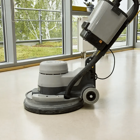A floor buffing machine moving around a hard floor by windows with a green grass view